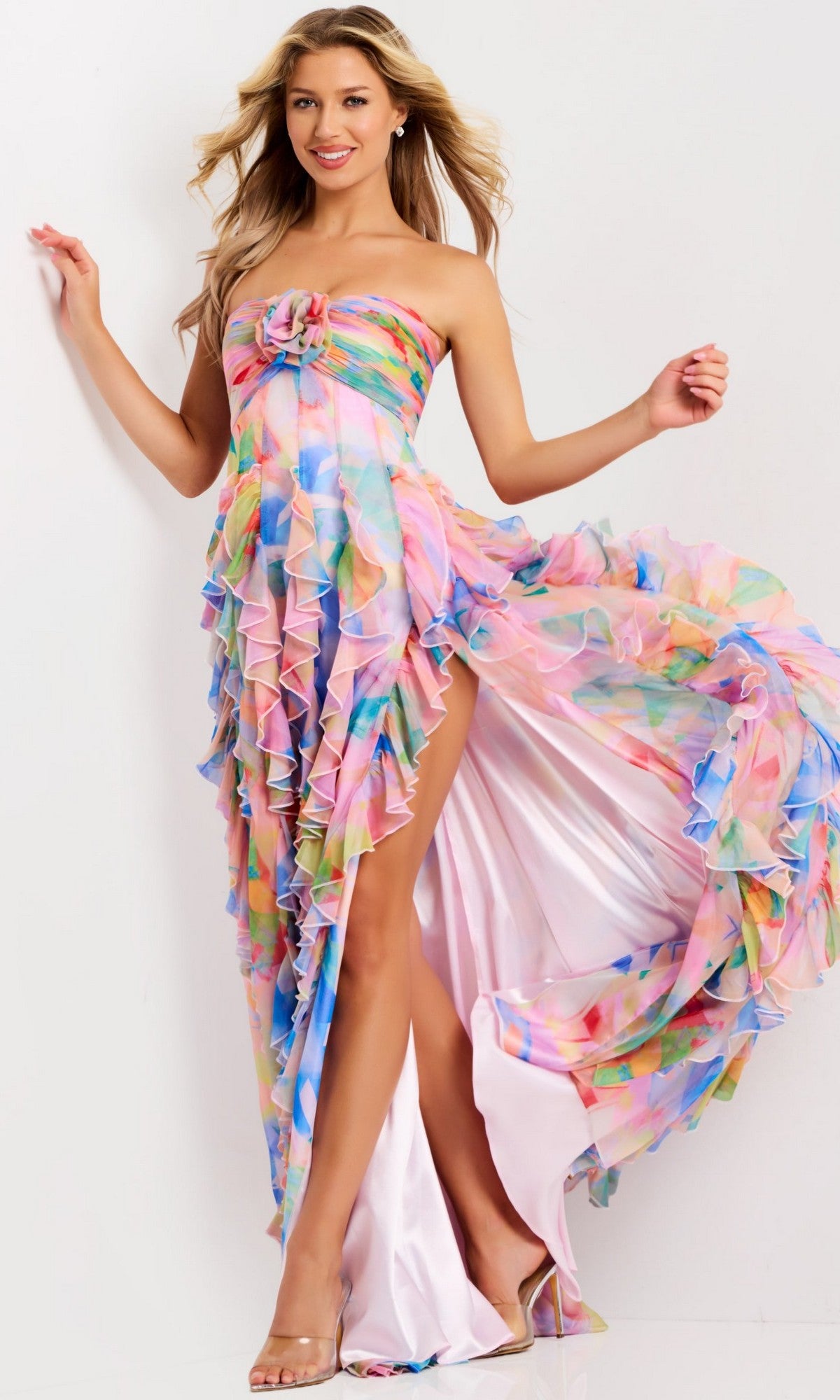 A woman poses against a white background, slightly lifting the skirt of the Jovani 46381 Long Prom Dress in a print color, featuring a strapless chiffon design with multicolored ruffles and a high slit.